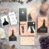 The Black Violet Tarot - Cherry Blossom Edition is displayed on a table with crystals and foliage. The minimalist black and pink designs evoke cherry blossoms, with elegant figures and symbols. The deck's box and guidebook are also in view.