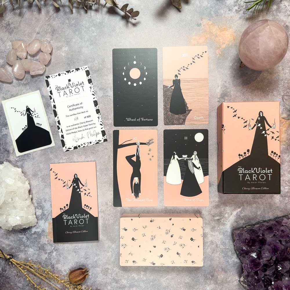 The Black Violet Tarot - Cherry Blossom Edition is displayed on a table with crystals and foliage. The minimalist black and pink designs evoke cherry blossoms, with elegant figures and symbols. The deck's box and guidebook are also in view.