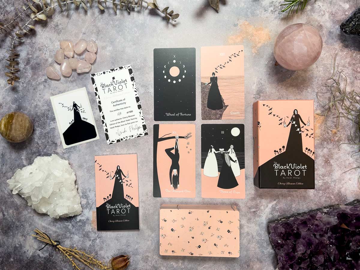 A flat lay of The Black Violet Tarot - Cherry Blossom Edition features black, white, and peach illustrations. Cards sit on a textured surface with decorative rose quartz, green branches, and crystals. A box is included.