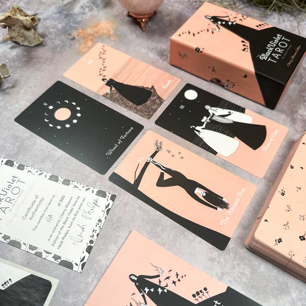 The Black Violet Tarot - Cherry Blossom Edition cards, with minimalist pink and black art depicting figures and celestial themes, rest on a textured surface. The deck's open box maintains the cherry blossom design.