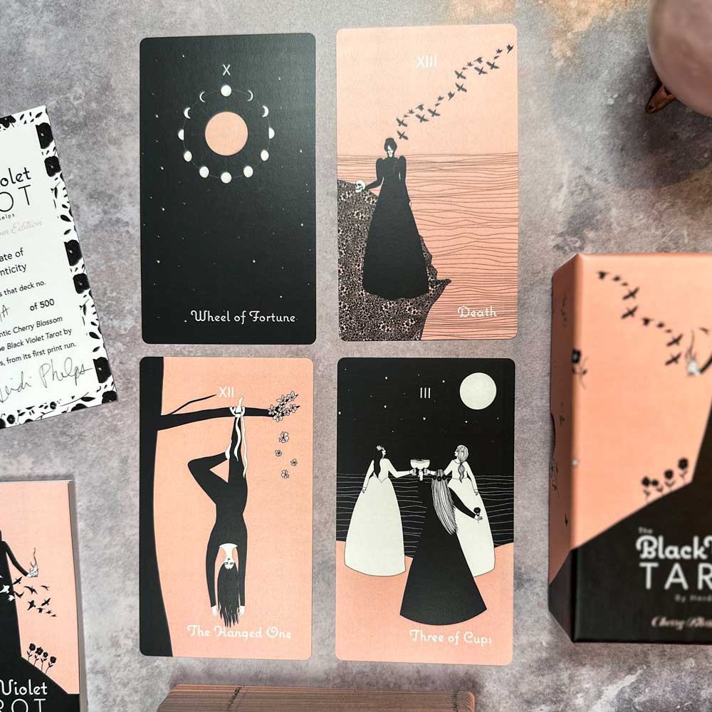 Four cards from The Black Violet Tarot - Cherry Blossom Edition are shown: Wheel of Fortune, Death, The Hanged One, and Three of Cups. They feature minimalist black and cherry blossom art. A tarot box is partially visible on the right.