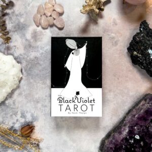 The Black Violet Tarot - Deluxe" box sits gracefully on a marble surface, surrounded by sparkling crystals like amethyst and rose quartz. The design features a stylized figure in flowing robes, embodying the deck's essence.