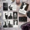 A set of tarot cards, The Black Violet Tarot - Deluxe, is laid out on a stone surface. The black and white illustrated cards include The Fool and The Magician. Crystals are also present.
