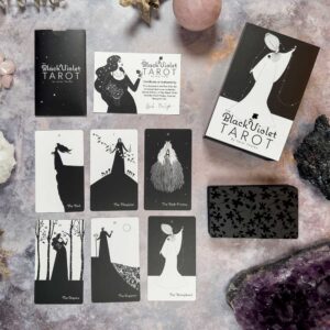 A set of tarot cards, The Black Violet Tarot - Deluxe, is laid out on a stone surface. The black and white illustrated cards include The Fool and The Magician. Crystals are also present.