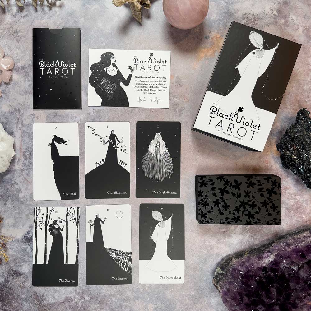 A set of tarot cards, The Black Violet Tarot - Deluxe, is laid out on a stone surface. The black and white illustrated cards include The Fool and The Magician. Crystals are also present.