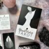 A box named "The Black Violet Tarot - Deluxe" sits on a textured surface, featuring a black and white illustration of someone in a long dress, with tarot cards, a pink stone sphere, and a dark rock surrounding it.
