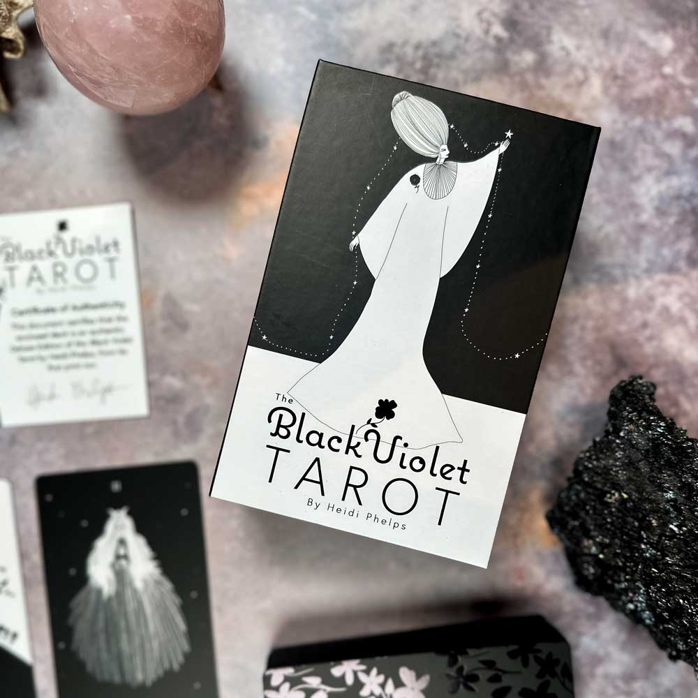 A box named "The Black Violet Tarot - Deluxe" sits on a textured surface, featuring a black and white illustration of someone in a long dress, with tarot cards, a pink stone sphere, and a dark rock surrounding it.