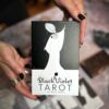 Hands adorned with rings and black nail polish hold The Black Violet Tarot - Deluxe box by Heidi Phelps, featuring a black-and-white illustration of a figure in a flowing gown reaching for a star, radiating enchantment.