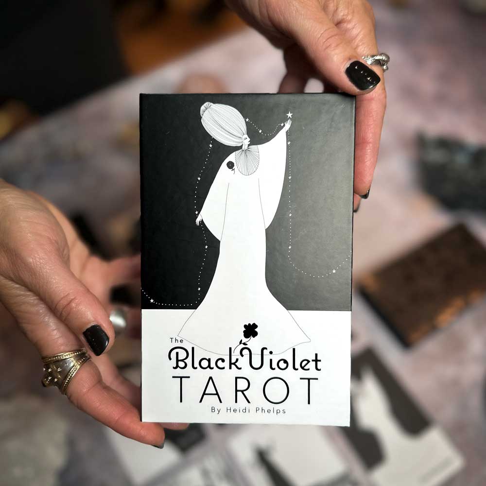Hands adorned with rings and black nail polish hold The Black Violet Tarot - Deluxe box by Heidi Phelps, featuring a black-and-white illustration of a figure in a flowing gown reaching for a star, radiating enchantment.