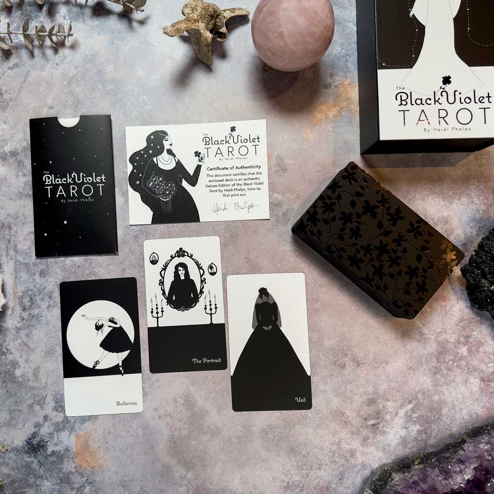 The Black Violet Tarot - Deluxe deck lies on a stone surface, with cards like "Celebration," "The Portrait," and "Void." Its black and white mystical design, highlighted by a pink sphere and leaf, adds to the enchanting scene.