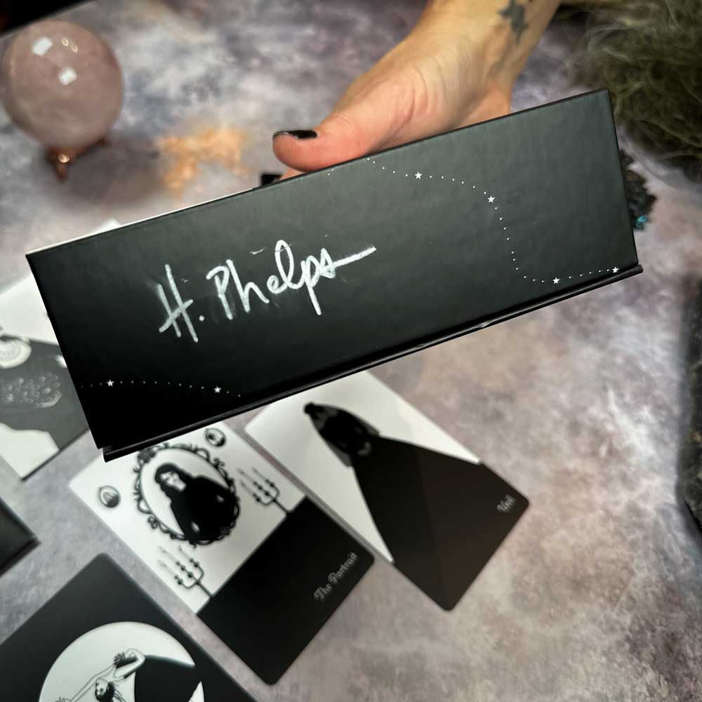 A hand holds a signed black box labeled "H. Phelps" from The Black Violet Tarot - Deluxe. Tarot cards featuring black and white designs are scattered on a textured gray surface, with a pink sphere providing an intriguing backdrop.