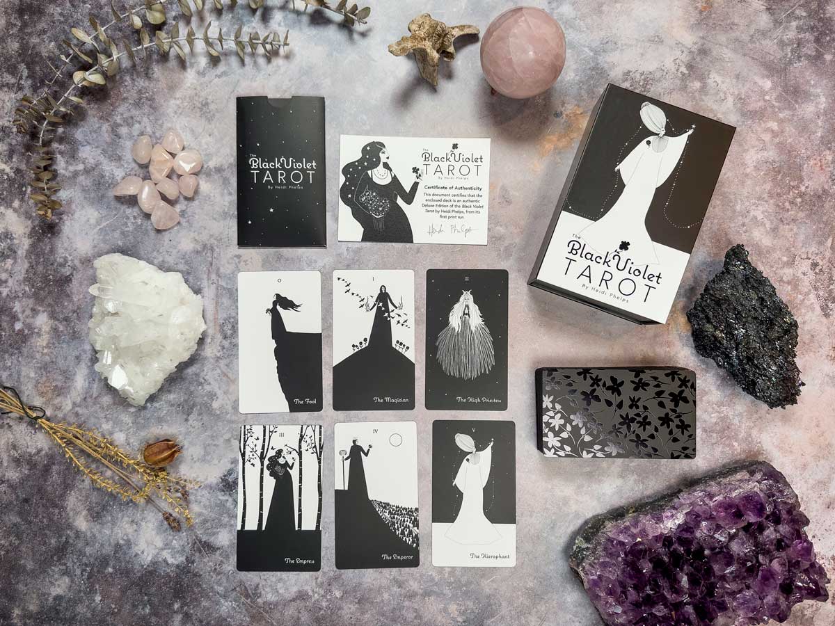 A deluxe tarot card set named "The Black Violet Tarot - Deluxe" is displayed with five cards, a guidebook, and the deck box, all surrounded by crystals, a pink sphere, and dried botanicals on a textured surface.