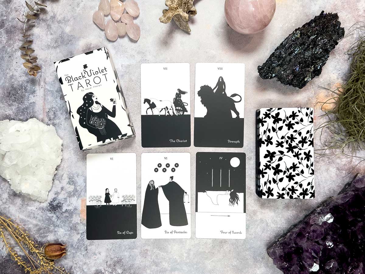 On a textured surface, a tarot spread from The Black Violet Tarot - The White Deck is surrounded by crystals and herbs, featuring The Chariot, Strength, 4 of Cups, 9 of Pentacles, and 4 of Wands.