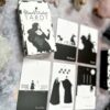 The Black Violet Tarot - The White Deck showcases exquisite black and white illustrations. Five cards spread out from the deck, surrounded by crystals on a patterned surface, complete this enchanting setup with its elegant box.