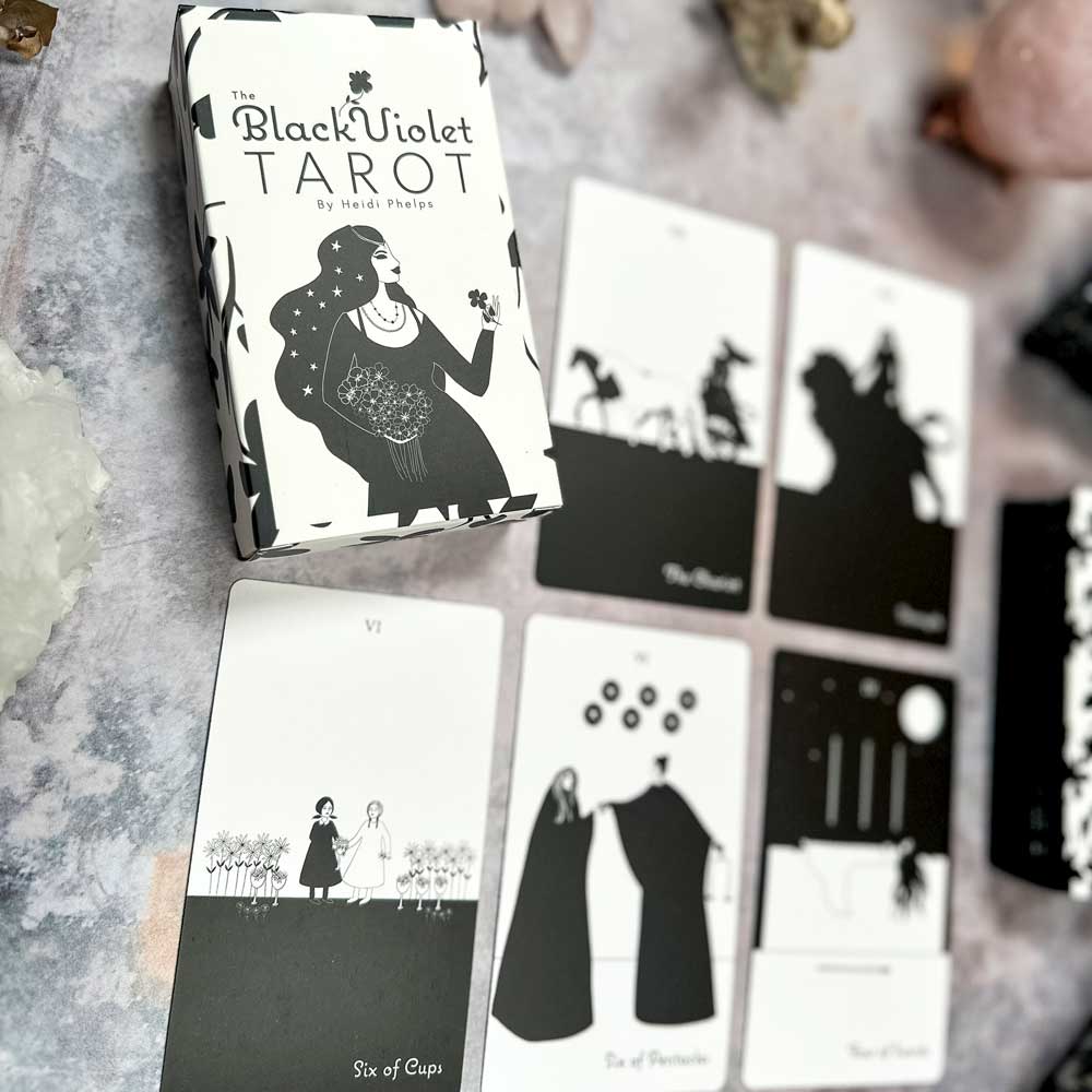 The Black Violet Tarot - The White Deck showcases exquisite black and white illustrations. Five cards spread out from the deck, surrounded by crystals on a patterned surface, complete this enchanting setup with its elegant box.