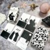 The Black Violet Tarot - The White Deck is elegantly showcased with six cards, featuring monochromatic illustrations and patterned backs. Surrounded by crystals and stones on a marble surface, it exudes mystical allure.