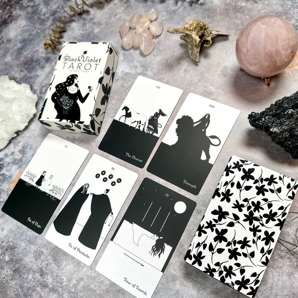 The Black Violet Tarot - The White Deck is elegantly showcased with six cards, featuring monochromatic illustrations and patterned backs. Surrounded by crystals and stones on a marble surface, it exudes mystical allure.