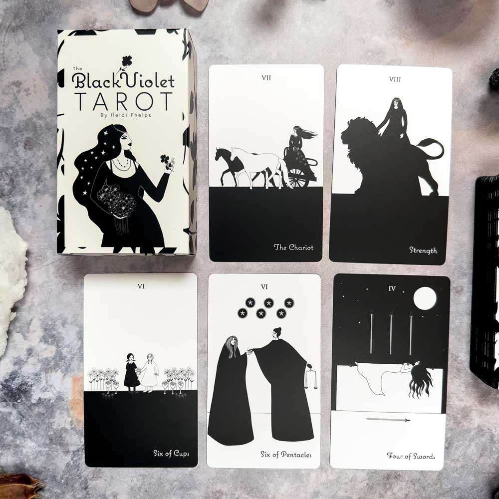 The Black Violet Tarot - The White Deck showcases five cards: The Chariot, Strength, Six of Cups, Six of Pentacles, and Four of Swords. Each card features black and white illustrations with diverse symbolic imagery.