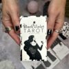Black-polished nails hold a box labeled "The Black Violet Tarot - The White Deck" by Heidi Phelps. Monochrome tones depict a woman with flowers, while tarot cards and crystals in the background enhance the mystical vibe.