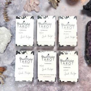 The Black Violet Tarot - The White Deck showcases six distinct boxes on a textured background with crystals and stones. Each box is signed and numbered, enhancing its unique appeal.