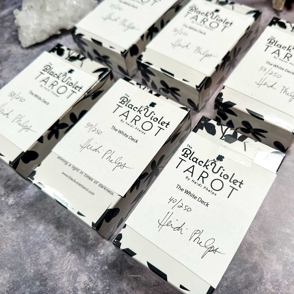 Several tarot boxes titled "The Black Violet Tarot - The White Deck" are arranged on a surface, each individually numbered and signed by Heidi Phelps. They feature a black and white floral design that elegantly enhances the sides.