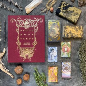 Flat lay of the "Blood Moon Tarot - Deluxe" set on textured ground, featuring the red and gold book, tarot cards, crystals, twigs, stones, a jawbone, and a black pouch with golden designs.