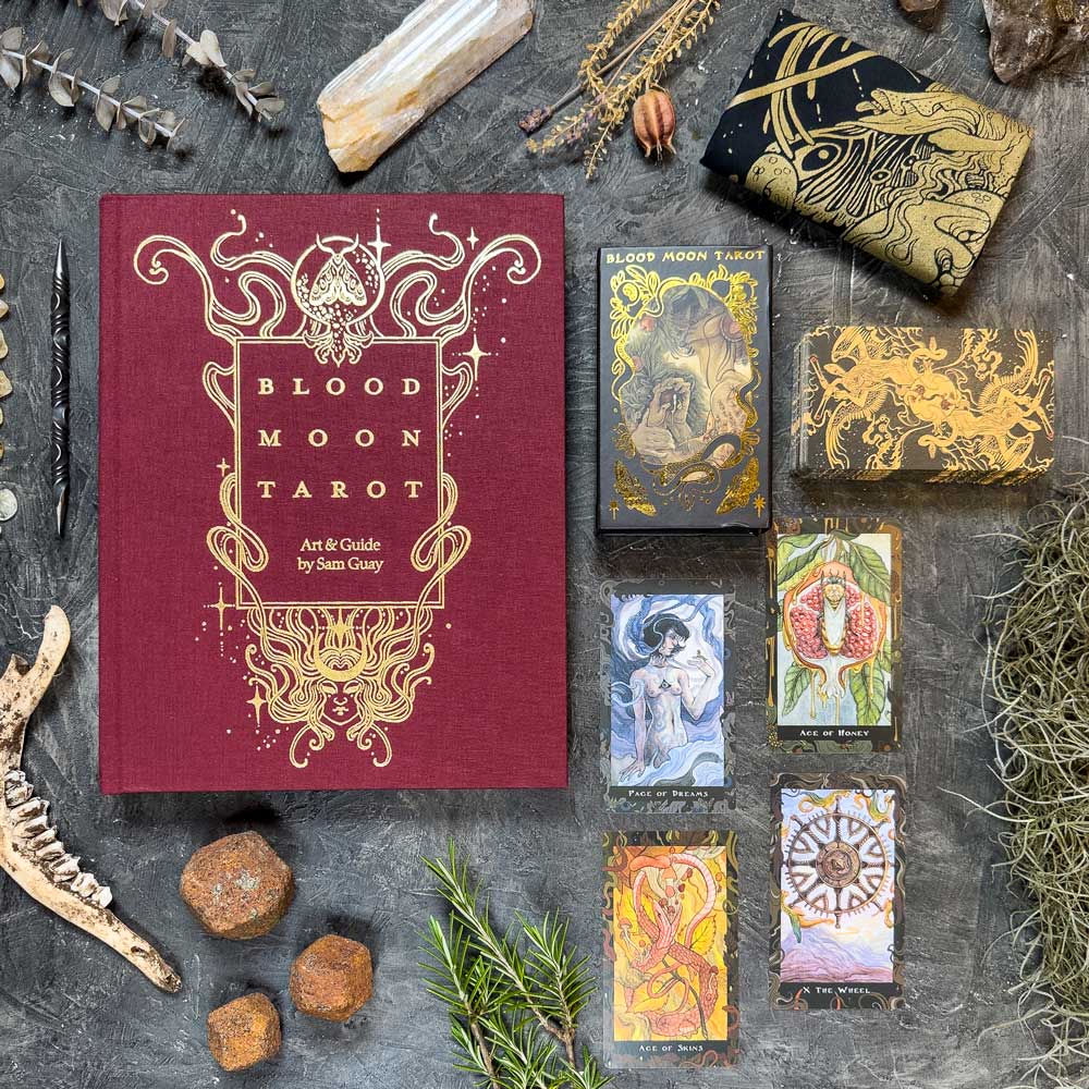 Flat lay of the "Blood Moon Tarot - Deluxe" set on textured ground, featuring the red and gold book, tarot cards, crystals, twigs, stones, a jawbone, and a black pouch with golden designs.