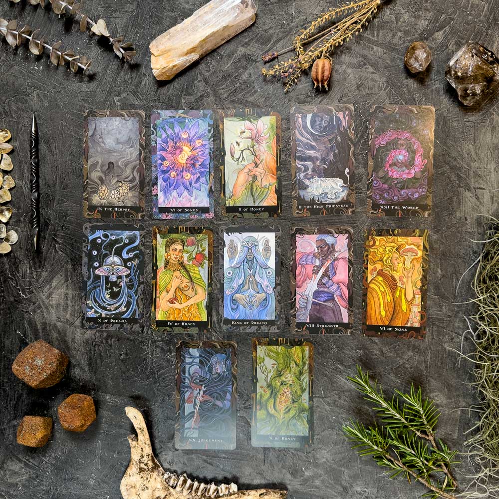 A set of 12 vibrant Blood Moon Tarot - Deluxe cards is elegantly displayed on a textured gray surface, surrounded by stones, herbs, a bone, and dried plants to create a mystical and earthy atmosphere.