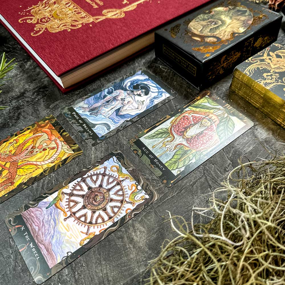 The Blood Moon Tarot - Deluxe cards lie on a textured surface by a red book and box, mesmerizing with their intricate designs. Sprigs of greenery and a decorative piece add to the aura.