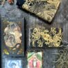 A set of Blood Moon Tarot - Deluxe cards rests on a stone surface, featuring intricate illustrations with gold and red accents. A gold and black cloth drapes the top left corner, with decorative elements and other cards peeking through to enhance the scene's allure.