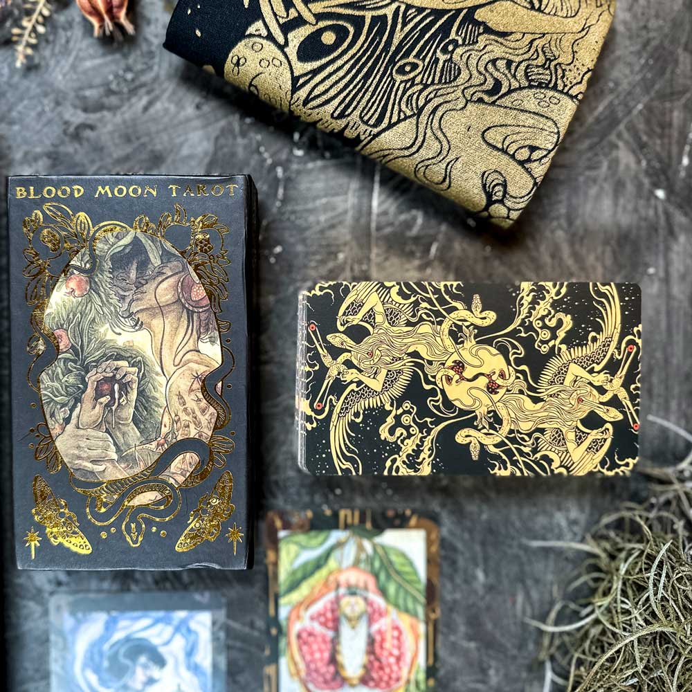 A set of Blood Moon Tarot - Deluxe cards rests on a stone surface, featuring intricate illustrations with gold and red accents. A gold and black cloth drapes the top left corner, with decorative elements and other cards peeking through to enhance the scene's allure.