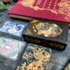 A black tarot card box adorned with golden lion designs is set on a dark surface, accompanied by three Blood Moon Tarot - Deluxe cards and an ornate red book. The cards showcase various artistic illustrations.
