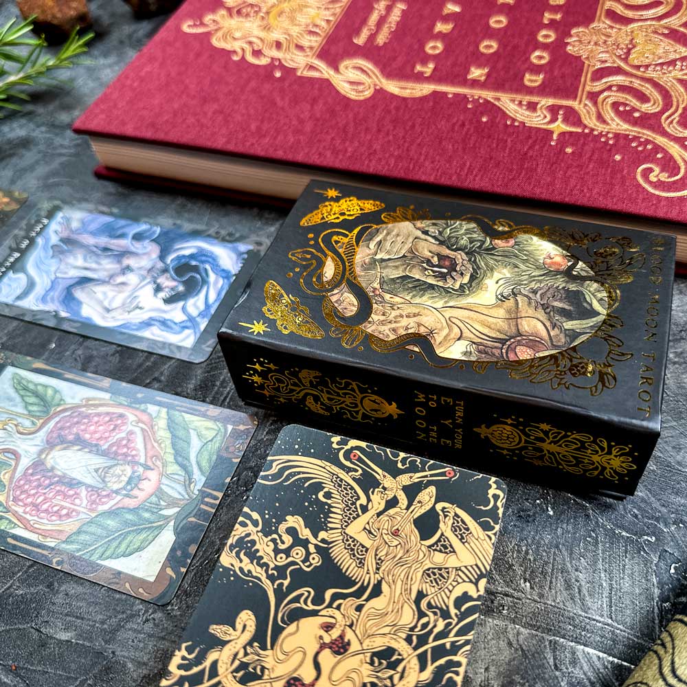 A black tarot card box adorned with golden lion designs is set on a dark surface, accompanied by three Blood Moon Tarot - Deluxe cards and an ornate red book. The cards showcase various artistic illustrations.