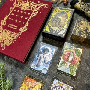 The red "Blood Moon Tarot - Deluxe" book lies open on a table alongside a tarot deck box with intricate gold designs. Several detailed, colorful tarot cards are spread out, and a sprig of greenery is nearby.