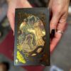 A close-up shows hands with dark nail polish and a silver ring holding the "Blood Moon Tarot - Deluxe" box. The box features intricate gold floral and leaf patterns with a mystical being at the center, enhancing the scene's enchantment.