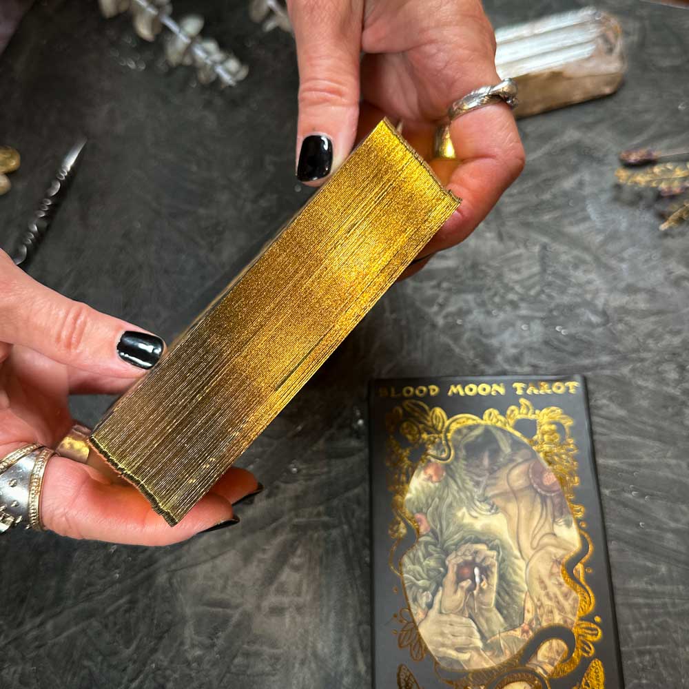 Black-polished, ring-adorned hands hold the "Blood Moon Tarot - Deluxe" deck with gold-edged cards. A partially visible, intricately designed tarot card lies below, evoking mystery.