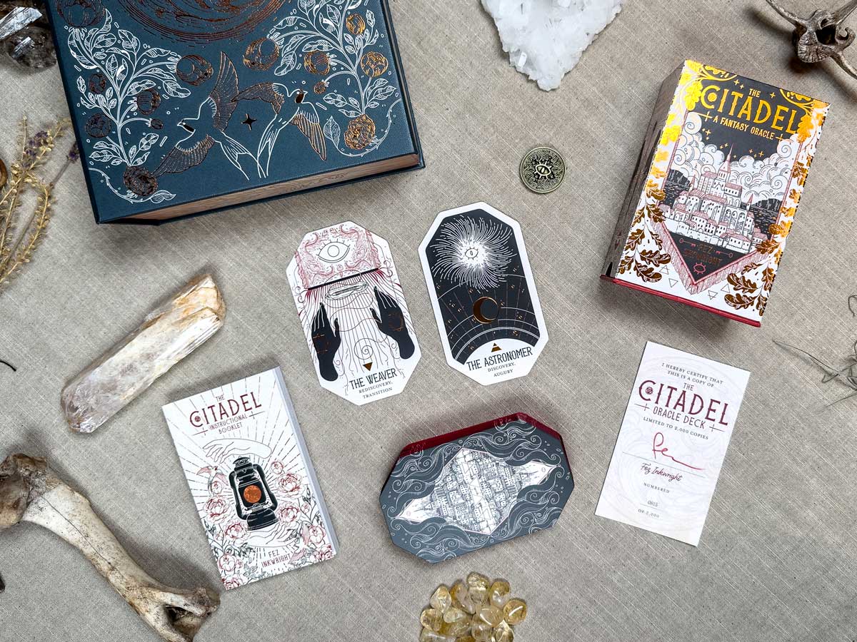 A flat lay of mystical items on fabric, featuring The Citadel Oracle - LE Boxed Set. It includes a pair of oracle cards, ornate boxes, a rune-like piece, crystals, a bone, and a parchment-style sheet labeled "Citadel," all in an ornate and mystical theme.