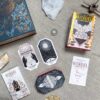 A flat lay displays The Citadel Oracle - LE Boxed Set, highlighting its fantasy oracle cards with celestial designs. Nearby, crystals and a metallic coin rest on a textured surface, creating an enchanting scene.