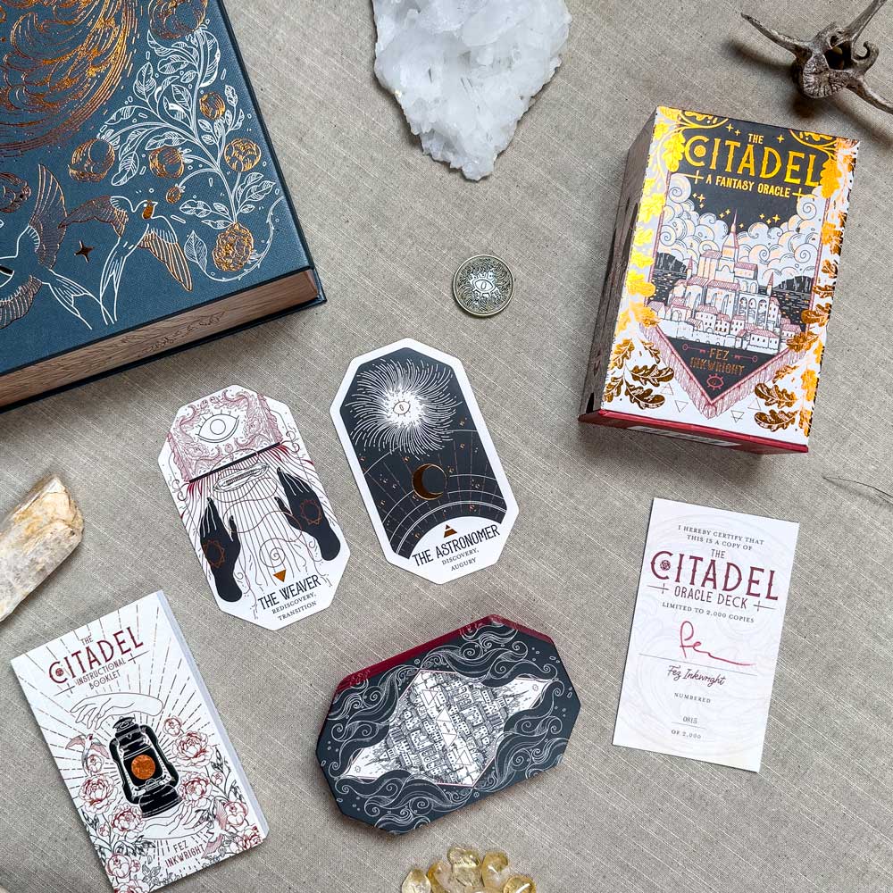 A flat lay displays The Citadel Oracle - LE Boxed Set, highlighting its fantasy oracle cards with celestial designs. Nearby, crystals and a metallic coin rest on a textured surface, creating an enchanting scene.