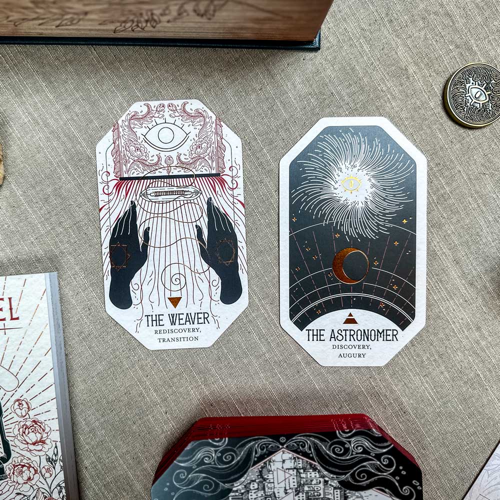 Two oracle cards from The Citadel Oracle - LE Boxed Set lie on fabric: "The Weaver," showing a figure weaving under an eye, and "The Astronomer," illustrating a celestial sun, planets, and stars. A coin and book corner are visible nearby.