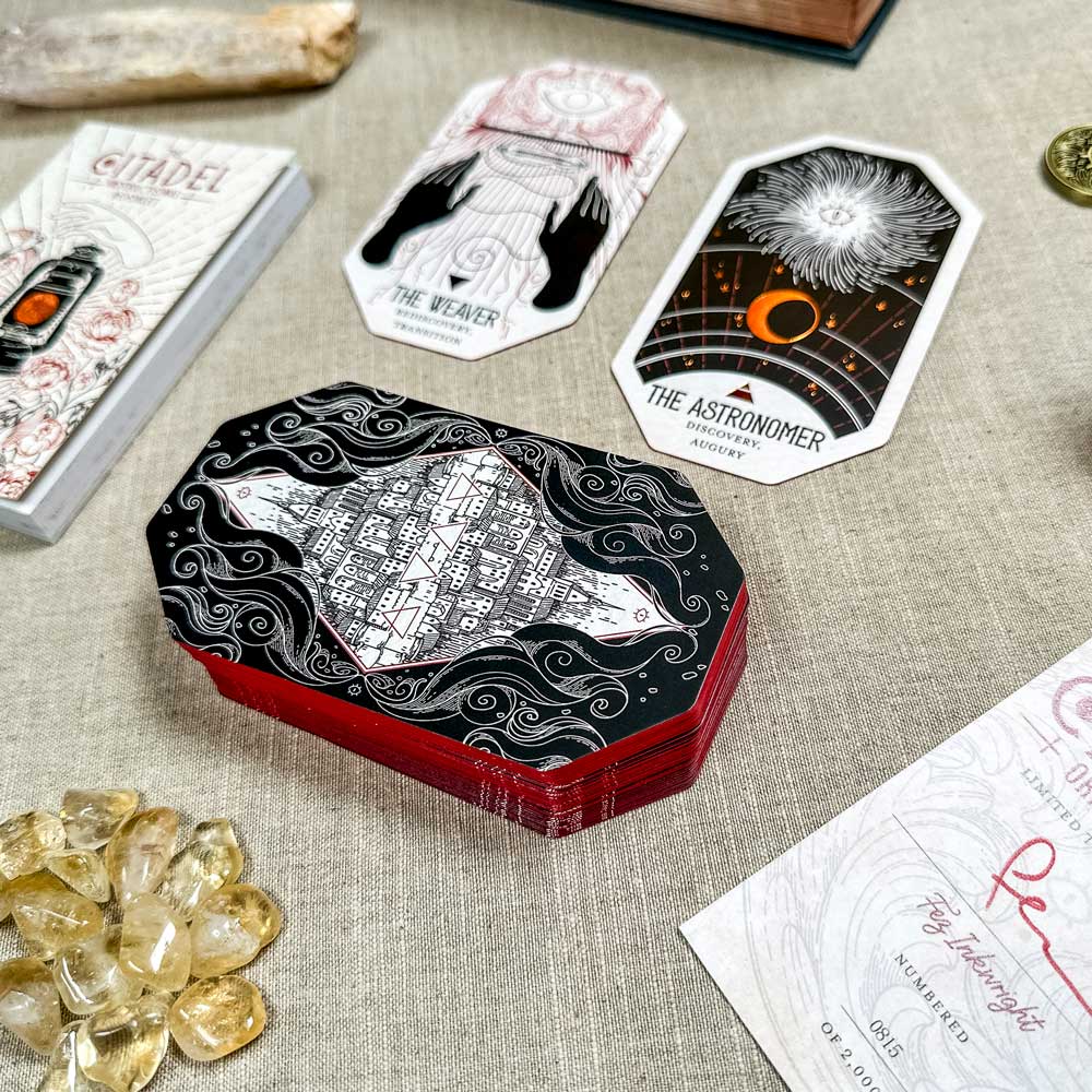 A spread of oracle cards from The Citadel Oracle - LE Boxed Set, featuring "The Weaver" and "The Astronomer," is beautifully displayed among crystals and mystical items on a textured beige table.