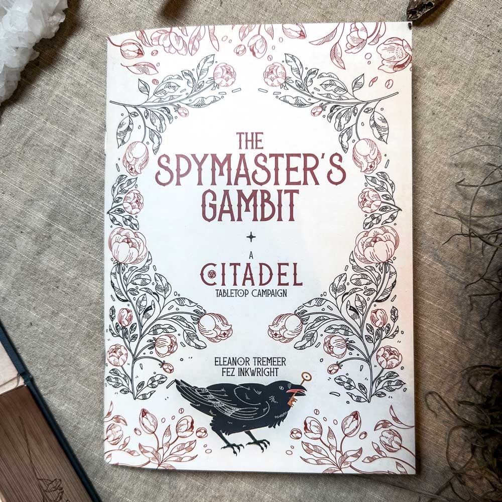 Cover of "The Spymaster's Gambit + A Citadel Tabletop Campaign" by Eleanor Tremeer and Fez Inkwright features ornate floral and vine designs with a black bird at the bottom, reminiscent of The Citadel Oracle - LE Boxed Set. Placed on a textured surface.