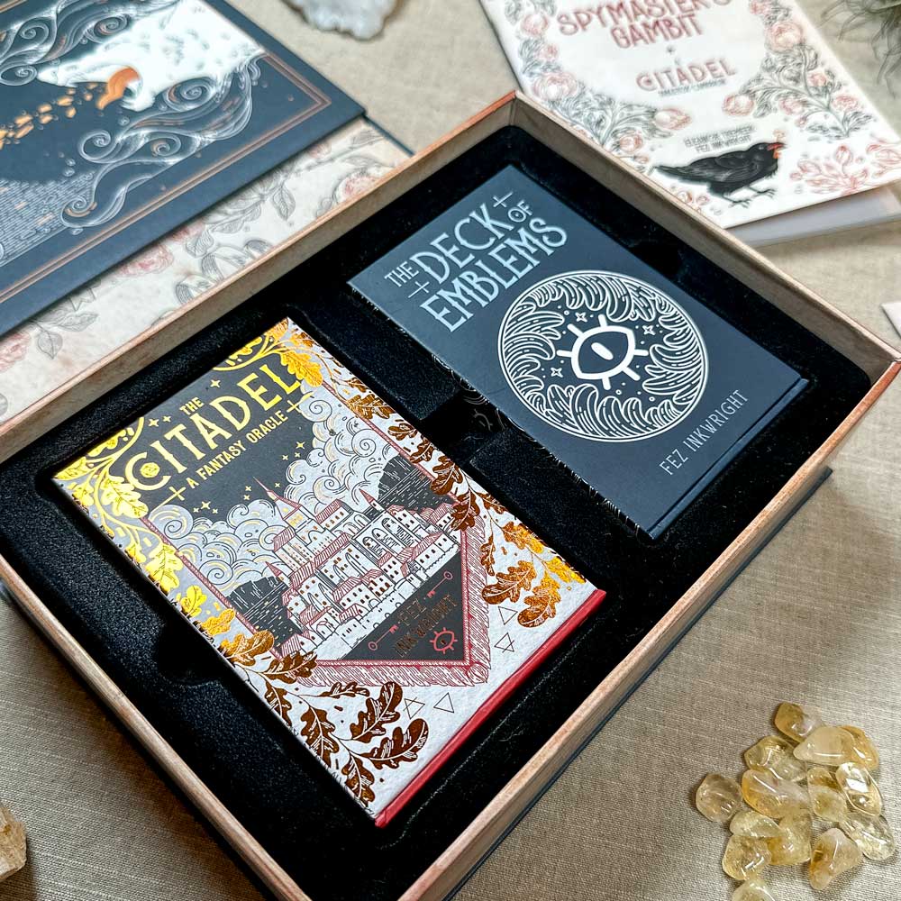 The Citadel Oracle - LE Boxed Set includes two captivating decks: "The Citadel: A Fantasy Racer" with a vibrant castle design and "The Deck of Emblems" featuring a circular motif, displayed open on a table amidst scattered gemstones.