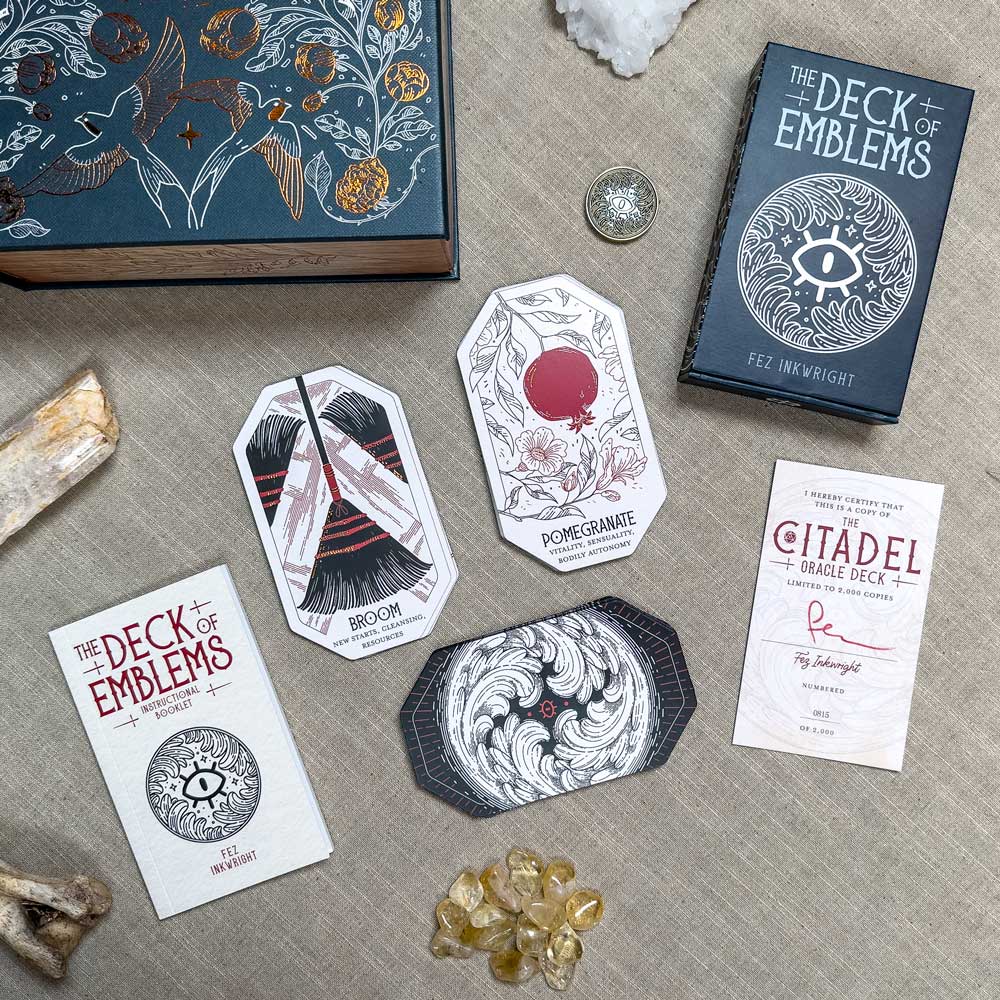 The Citadel Oracle - LE Boxed Set is displayed on a textured surface, featuring intricately designed oracle cards with motifs like a broom and pomegranate. A box labeled "The Deck of Emblems" and various stones accompany the collection.