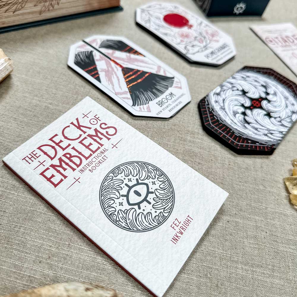 Close-up of "The Deck of Emblems" card game on a textured surface. The scene highlights the instruction booklet with a circular emblem similar to "The Citadel Oracle - LE Boxed Set," surrounded by intricately patterned and symbolized cards.