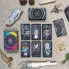 The Cosmic Visions Tarot set, vibrant and intricately designed, is showcased alongside crystals, a white sage bundle, and natural elements like rocks and twigs. All items are elegantly spread over a beige fabric surface.