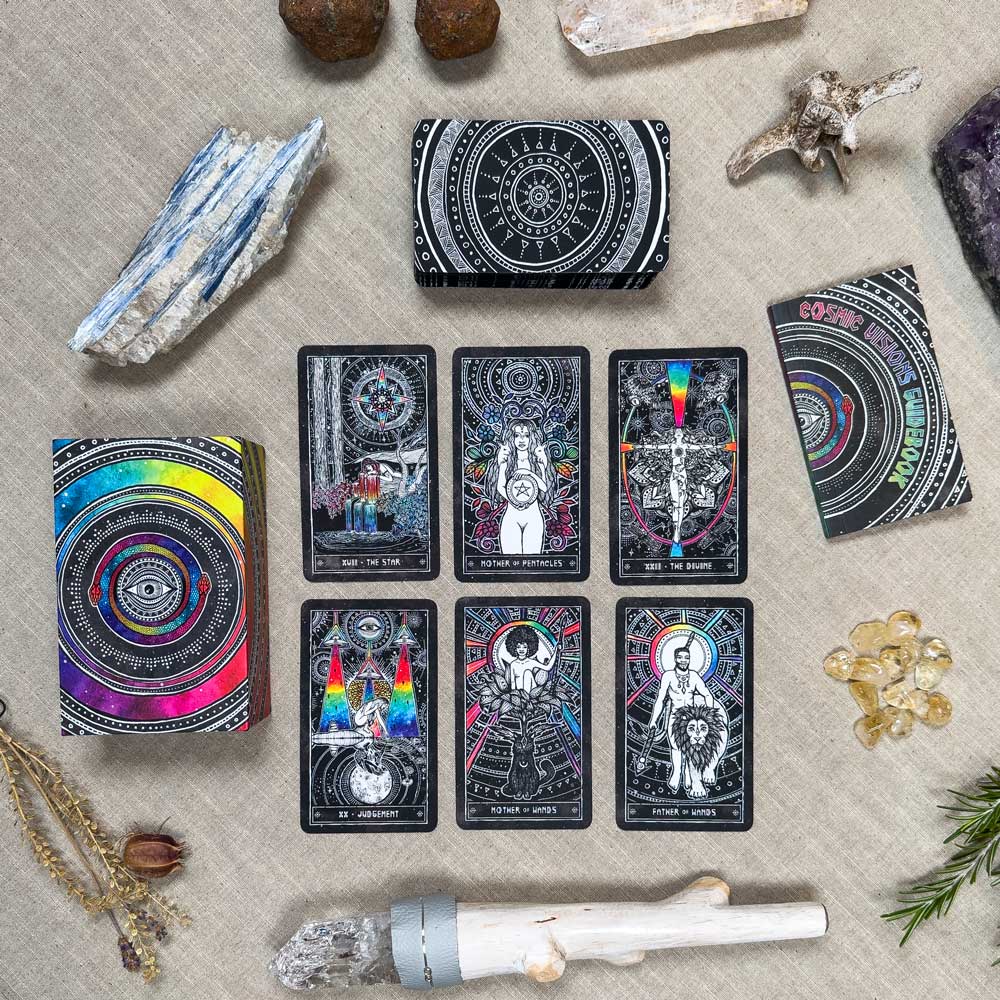 The Cosmic Visions Tarot set, vibrant and intricately designed, is showcased alongside crystals, a white sage bundle, and natural elements like rocks and twigs. All items are elegantly spread over a beige fabric surface.
