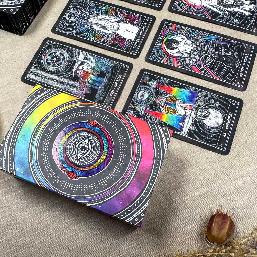 The Cosmic Visions Tarot deck and box with vibrant, intricate designs are displayed on a beige surface. The cards showcase detailed mystical illustrations, accompanied by a beetle and dried plants that amplify their enchanting aura.