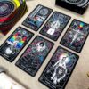 A set of six Cosmic Visions Tarot cards with intricate designs and vivid colors on dark backgrounds is laid out on a beige surface. A box with matching artwork is partially visible in the corner.