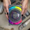 Hands holding a colorful Cosmic Visions Tarot notebook with concentric circles, dot patterns, and vibrant hues like blue, purple, red, and yellow centered around an eye design.